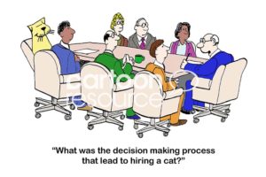 Color interview cartoon showing a smiling cat sitting in a work meeting.  The boss asks the employees around the meeting table, "what was the decision making process that led to hiring a cat?".