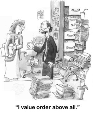 Office b&w cartoon showing a man with an extremely cluttered office. He says to a female coworker, "I value order above all".
