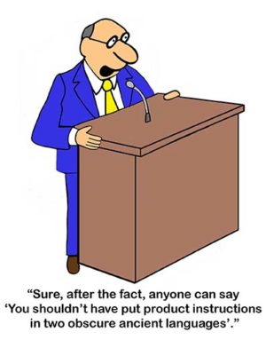 Office color cartoon showing a businessman standing and talking at a lectern stating, 'Sure, after the fact, anyone can say, "you shouldn't have put product instructions in two obscure ancient languages"'.