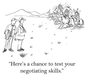 B&W, single-panel cartoon showing two professional men on the canyon lands, Indians are about to attack. One says this is a good time to practice 'negotiating skills'.